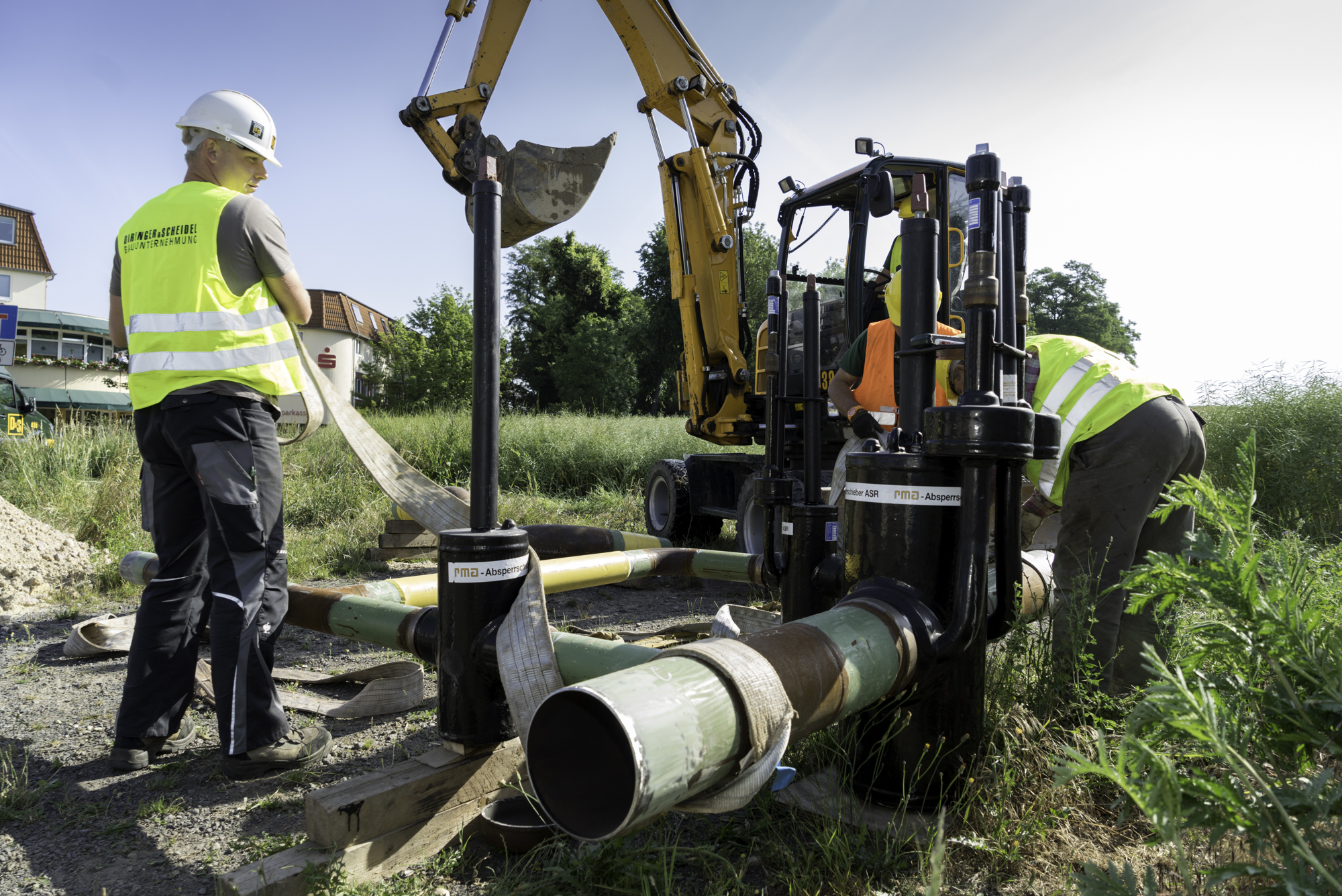 Drei Mitarbeiter und ein Bagger bei Arbeiten an Rohrleitungen auf einer Wiese